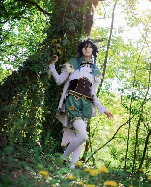 Nữ cosplayer đứng giữa rừng xanh trong tạo hình Venti, áo choàng lượn sóng nổi bật dưới ánh sáng tự nhiên nhẹ qua tán lá