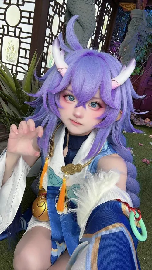 Ảnh Cosplay Honkai Star Rail với cosplayer tóc tím đeo tai thú, giơ tay chào nhẹ giữa nền cây xanh mờ phía sau.