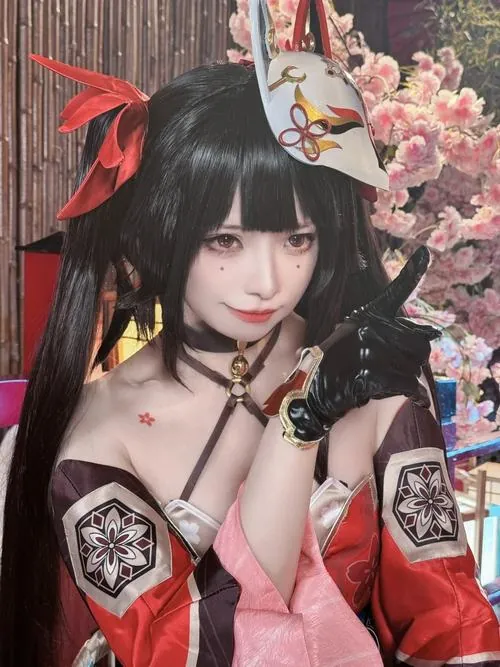 Cosplayer tóc đen trong bộ đồ đỏ đen ngồi ngoài vườn, biểu cảm dễ thương khiến khung ảnh trở nên gần gũi.