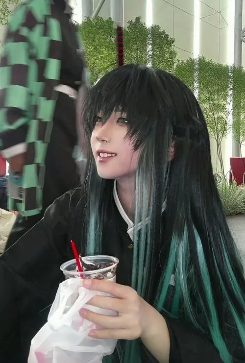 Nữ cosplayer ngồi ngoài trời bên ly nước, mái tóc xanh ngọc rủ qua vai và nụ cười nhẹ làm ảnh mềm hơn